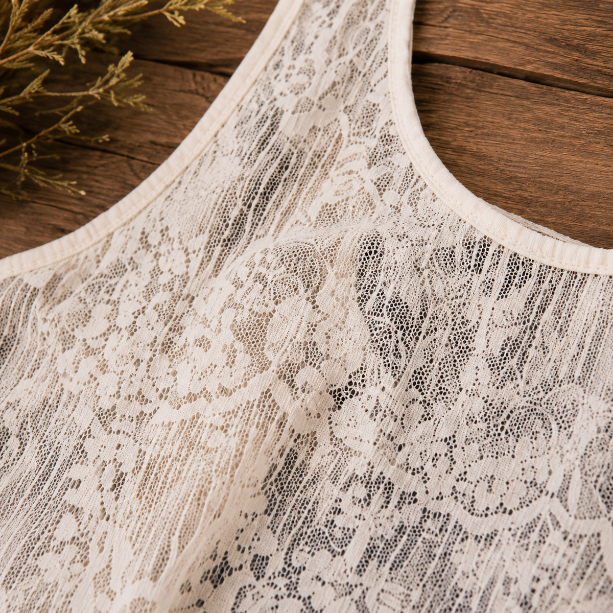 Close-up of a cream lace sleeveless top or dress laid flat on a rustic wooden surface, with dried floral stems in the corner.