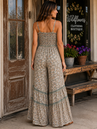 A woman stands outdoors in front of a rustic storefront wearing a sleeveless, wide-leg jumpsuit with a small floral print. The jumpsuit has thin straps, a smocked bodice, and tiered flared lower sections with teal-green trim.