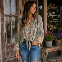 A woman stands on a rustic porch wearing a long-sleeve tie-front blouse with a muted floral print and green patchwork sleeves, paired with blue jeans. The background shows weathered wood doors and a window with the sign 'Wildflowers Clothing Boutique'.