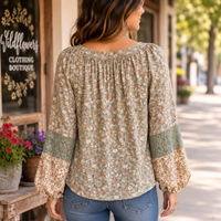 A woman viewed from behind wearing a long-sleeve blouse with a dense small floral print in muted green, beige, and orange tones, paired with blue jeans, standing on a sunny street in front of a boutique.