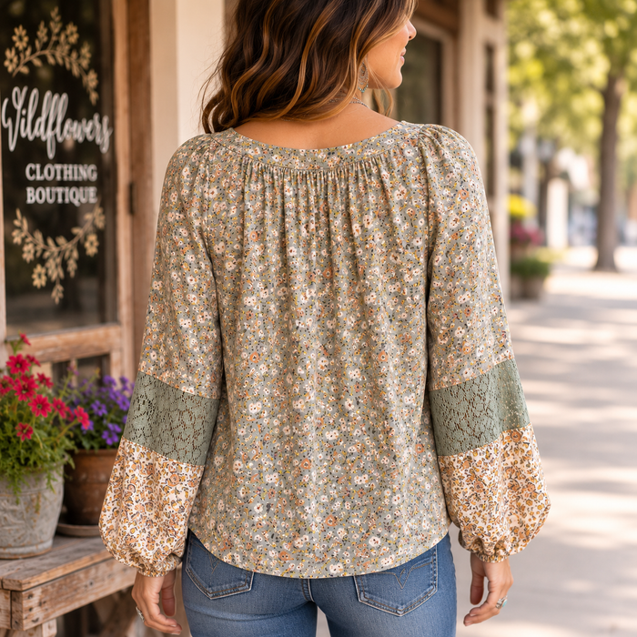 A woman viewed from behind wearing a long-sleeve blouse with a dense small floral print in muted green, beige, and orange tones, paired with blue jeans, standing on a sunny street in front of a boutique.