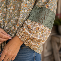 A close-up of a person wearing a long-sleeve blouse with a muted floral print, a solid sage green lace panel on the upper arm, and a matching floral lace cuff, paired with blue jeans.