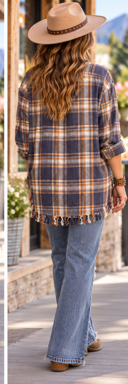 A woman shown from behind wearing a navy, orange, and beige plaid flannel shirt with fringe trim, blue jeans, and a tan wide-brim hat, standing outdoors on a sidewalk.