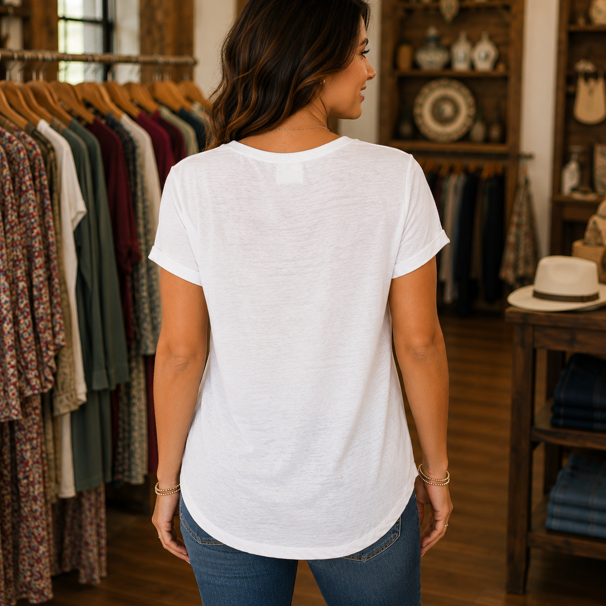 A woman viewed from behind wearing a white short-sleeve t-shirt and blue jeans inside a clothing boutique, with racks of garments and wooden shelving visible in the background.