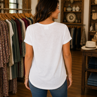 A woman viewed from behind wearing a white short-sleeve t-shirt and blue jeans inside a clothing boutique, with racks of garments and wooden shelving visible in the background.