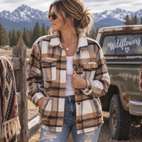 A woman wearing a tan, brown, and black plaid flannel overshirt over a white tank top and distressed blue jeans, standing outdoors in front of a rustic green pickup truck and snowy mountains.