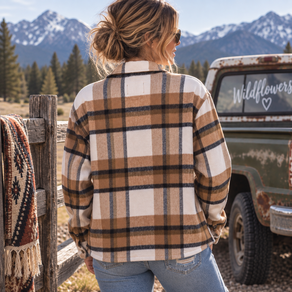 A woman viewed from behind wearing a tan, cream, and black plaid flannel shirt in an outdoor mountain setting next to a rustic green truck with a 'Wildflowers' decal.