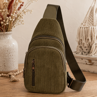 An olive green corduroy sling bag with a matching adjustable shoulder strap, displayed on a wooden surface with boho-style decorative props including a ceramic vase with dried flowers and a macrame wall hanging.