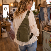 A woman in a rustic shop wearing an olive green corduroy sling backpack, viewed from behind as she looks over her shoulder.