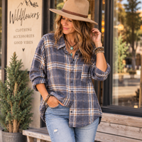 A woman wearing a blue and beige plaid button-down shirt, light blue jeans, and a tan wide-brim hat, standing outdoors in front of a storefront with a sign that reads 'Wildflowers'.