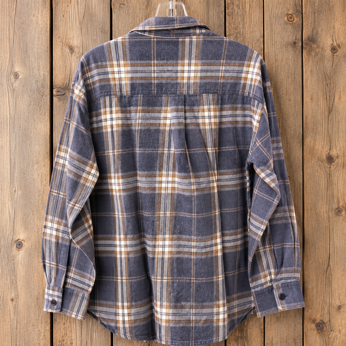 A long-sleeve plaid flannel shirt hanging on a hanger against a rustic wooden plank background. The shirt features a muted slate blue base with intersecting tan and white plaid lines.