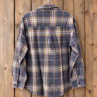 A long-sleeve plaid flannel shirt hanging on a hanger against a rustic wooden plank background. The shirt features a muted slate blue base with intersecting tan and white plaid lines.