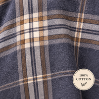A close-up of a plaid fabric with a textured weave, featuring navy blue, beige, brown, and white stripes. A circular badge in the bottom right corner reads '100% COTTON' with a small leaf illustration.