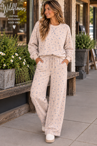 A woman models a matching two-piece lounge set consisting of a long-sleeve sweatshirt and wide-leg pants, both in a light cream/beige color with a small blue floral pattern. She is standing on a sidewalk in front of a storefront with potted plants.
