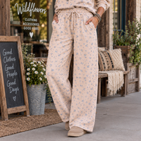 A person wearing light beige drawstring lounge pants with a small blue floral print, standing on a storefront porch. The scene includes a chalkboard sign reading 'Wildflowers Clothing Accessories Good Vibes,' potted flowers, a bench with pillows, and the person's hands in the pockets.