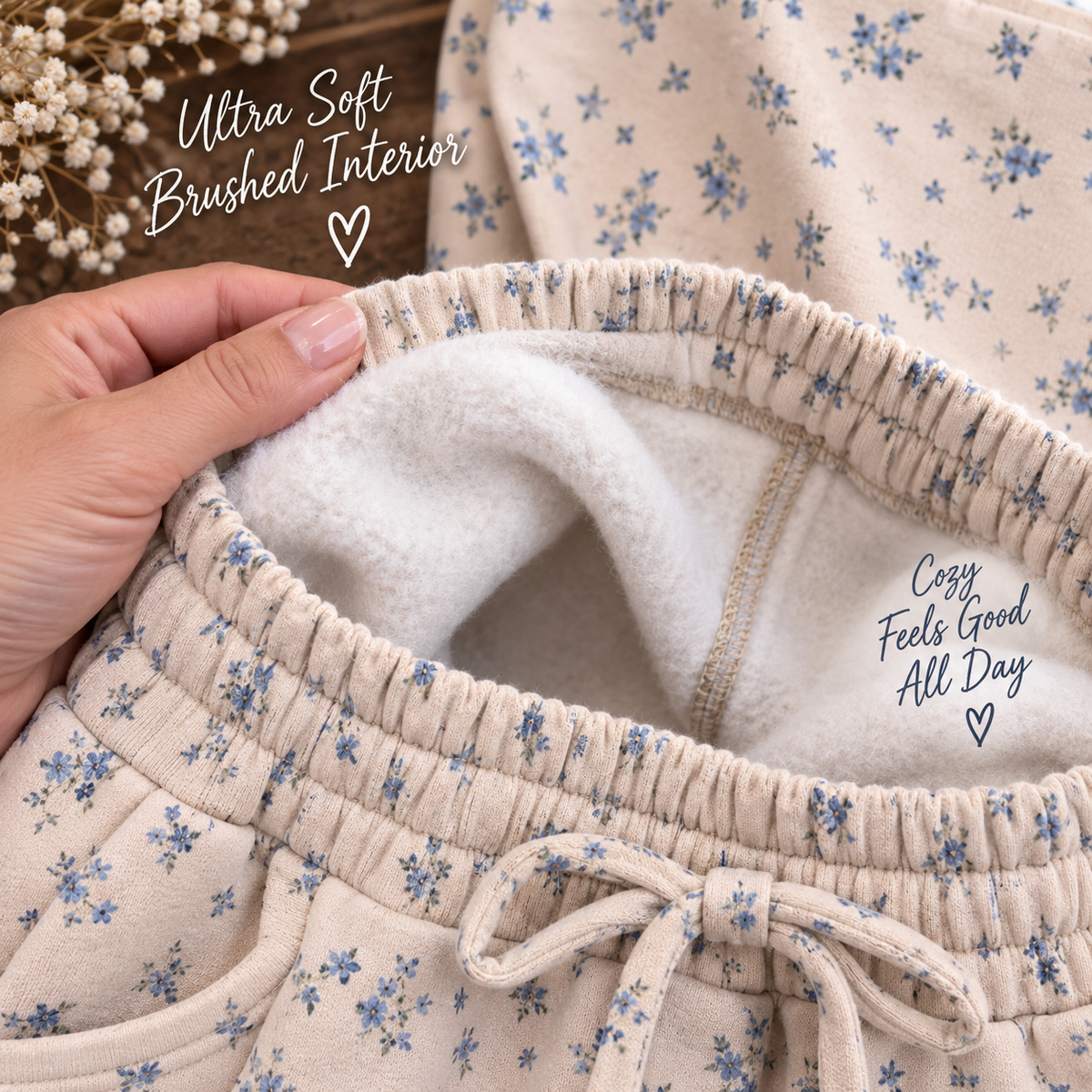 Close-up of a person's hand holding the waistband of cream-colored floral lounge pants to show the soft brushed interior lining. The pants feature a small blue floral print, elastic drawstring waist, and cozy fleece-like inner lining with printed text overlays.
