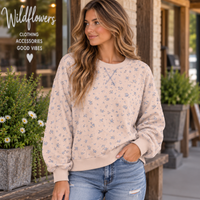 A woman wearing a light beige crewneck sweatshirt with a small blue floral pattern, paired with blue jeans, standing outdoors in front of a storefront with potted plants.