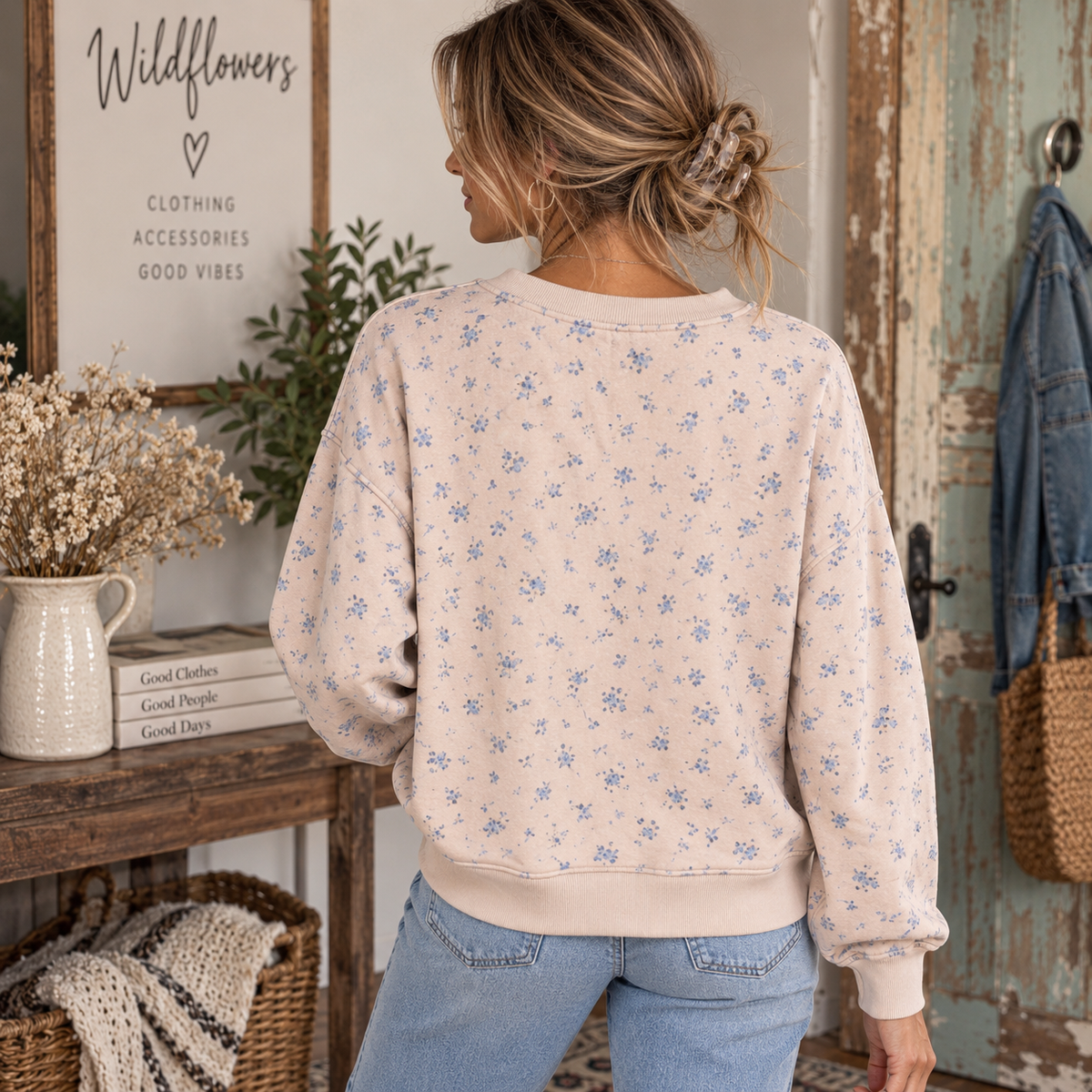 A woman viewed from behind wearing a cream-colored crewneck sweatshirt with a small blue floral pattern, paired with light blue jeans in a rustic, cozy room setting.