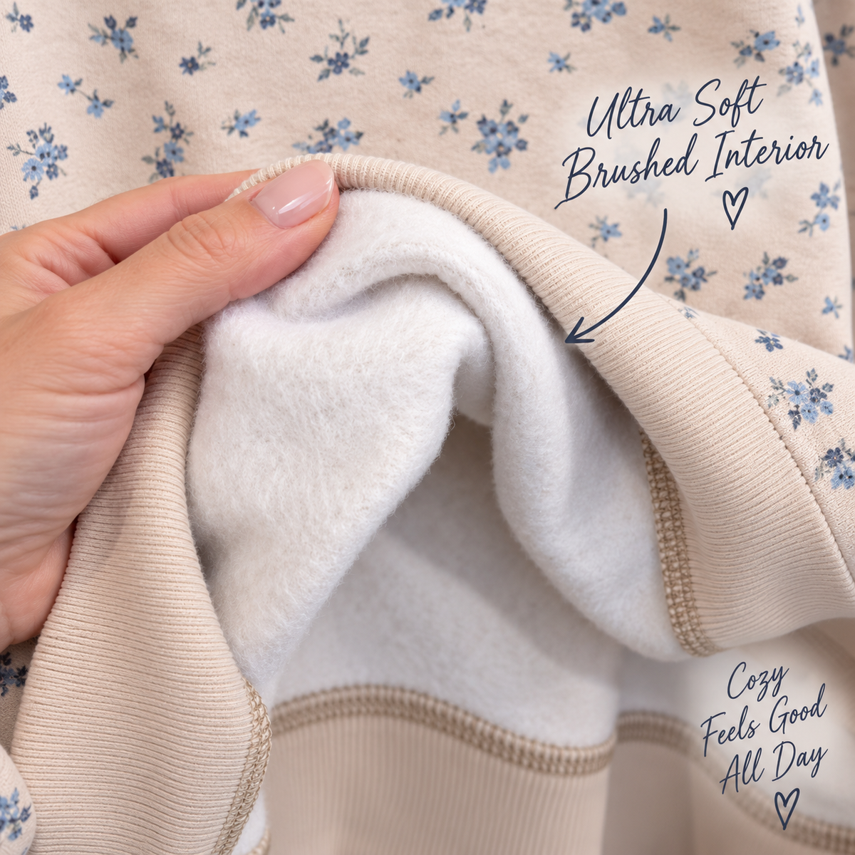 Close-up of a person's hand holding open a beige sweatshirt to show its ultra-soft brushed white interior lining, with blue floral print visible on the exterior. Text overlays highlight the 'Ultra Soft Brushed Interior' and 'Cozy Feels Good All Day'.