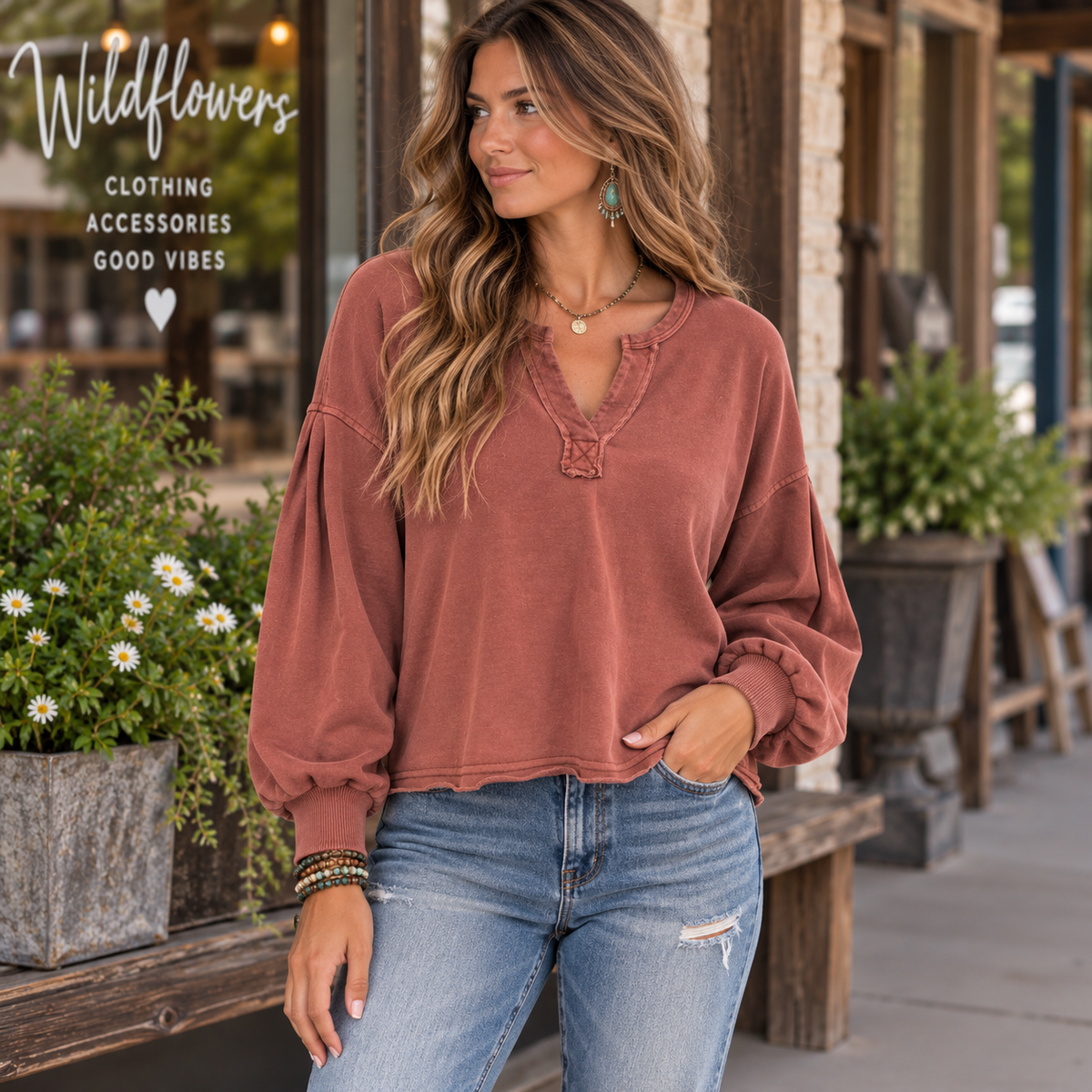 A woman wearing a dusty rose long-sleeve top with balloon sleeves and a split neckline, paired with blue jeans, standing outdoors in front of a storefront with plants and a bench. The Wildflowers logo is visible in the top left corner.