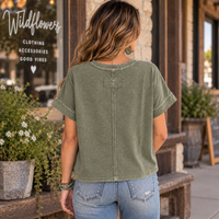 A woman viewed from behind wearing an olive green short-sleeve crop top with a central seam and decorative stitching, paired with light blue jeans, standing on a storefront porch.