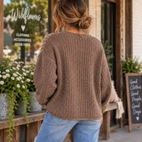 A woman viewed from behind wearing a brown chunky knit sweater and light blue jeans, standing on a storefront porch with plants and a chalkboard sign visible.
