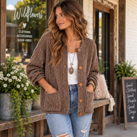 A woman stands on a storefront porch wearing a chunky brown knit cardigan with wooden buttons over a white top and light blue jeans. The cardigan has a textured cable-knit pattern and front pockets.