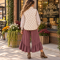A woman stands on a sidewalk in front of a storefront, shown from the back. She is wearing a sleeveless cream vest with a small tan floral pattern over a light beige long-sleeve top, paired with dusty rose wide-leg ruffled pants and brown ankle boots.