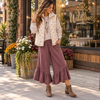 A woman stands on a sidewalk in front of a storefront wearing a cream vest with a floral pattern over a light top, mauve wide-leg ruffled pants, and brown ankle boots. The storefront has a 'Wildflowers Boutique' sign.