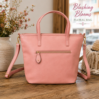 A dusty pink faux-leather tote bag with a front zip pocket and a matching shoulder strap, styled on a wooden table with a vase of flowers and a straw hat in the background. The text 'Blushing Blooms FLORAL BAG' is visible in the top right corner.
