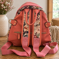 A coral pink canvas backpack with floral printed fabric panels and adjustable shoulder straps, shown from the back on a wooden surface with decorative home items in the background.