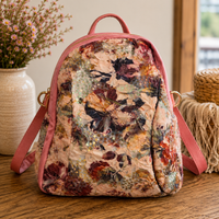 A small backpack with a pink leather trim and handle, featuring a vintage floral print with sequins on the front, displayed on a wooden table next to a vase of flowers.