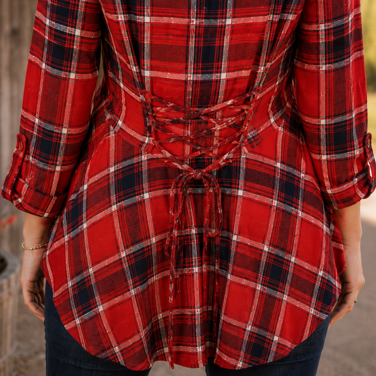 A red plaid long-sleeve top with a lace-up back detail, shown on a model from the back. The shirt features a classic tartan pattern in red, navy, white, and black.