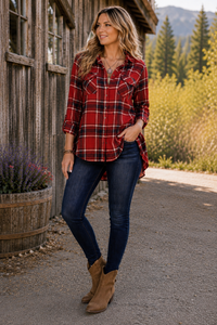 A woman stands outdoors in front of a rustic wooden building, wearing a red and black plaid button-up flannel shirt, dark blue skinny jeans, and tan ankle boots.