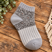 A single ankle sock with a gray and black marled knit pattern and solid gray ribbed sections, displayed on a rustic wooden surface with decorative green leaves and a cream macrame fringe prop.