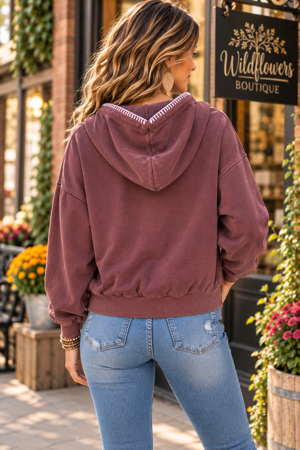 A woman viewed from behind wearing a dusty rose hooded sweatshirt and light blue jeans, standing outdoors in front of a storefront with a sign that reads Wildflowers Boutique.