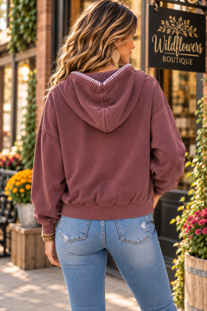 A woman viewed from behind wearing a dusty rose hooded sweatshirt and light blue jeans, standing outdoors in front of a storefront with a sign that reads Wildflowers Boutique.