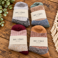 Four pairs of mo:vint New York knit socks arranged in a 2x2 grid on a rustic wooden surface. Each sock has a neutral paper band with the brand name 'mo:vint new york' printed on it.
