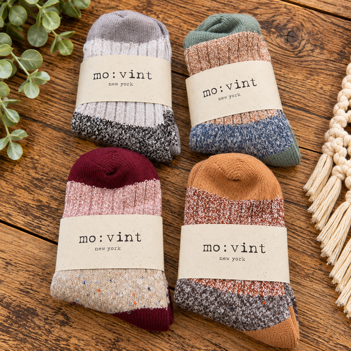 Four pairs of mo:vint New York knit socks arranged in a 2x2 grid on a rustic wooden surface. Each sock has a neutral paper band with the brand name 'mo:vint new york' printed on it.