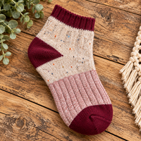 A single ankle sock laid flat on a rustic wooden surface, with green leaves and a cream woven textile partially visible at the edges. The sock has a cream upper with tiny multicolor speckles, a dark burgundy cuff and heel section, and a pale pink ribbed lower section with a burgundy toe.