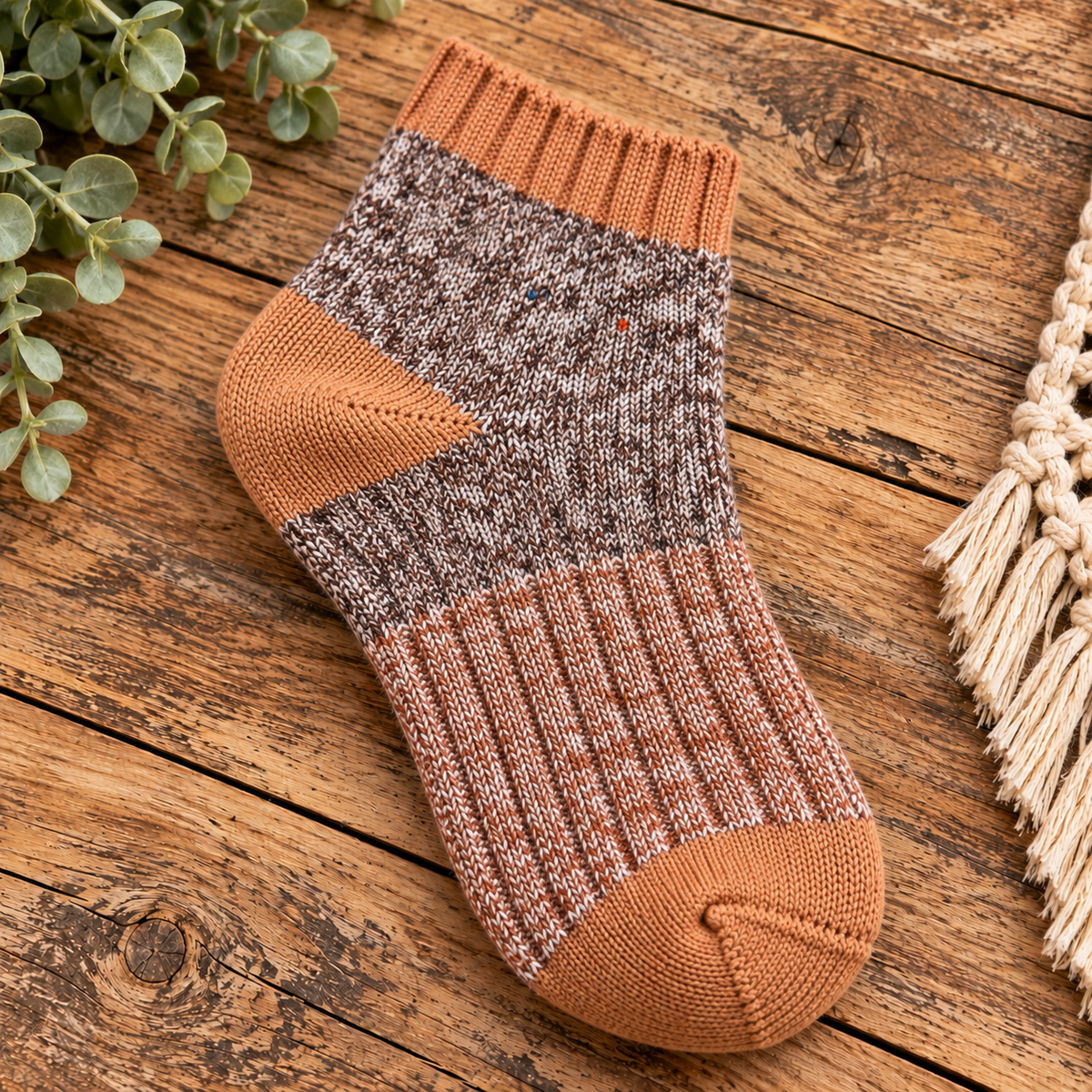 A single knit sock laid on a rustic wooden surface, with green leaves at the upper left and a cream fringed textile at the right. The sock has a heathered brown and cream body, rust orange ribbed cuff, heel, and toe, and a small embroidered red accent near the top.