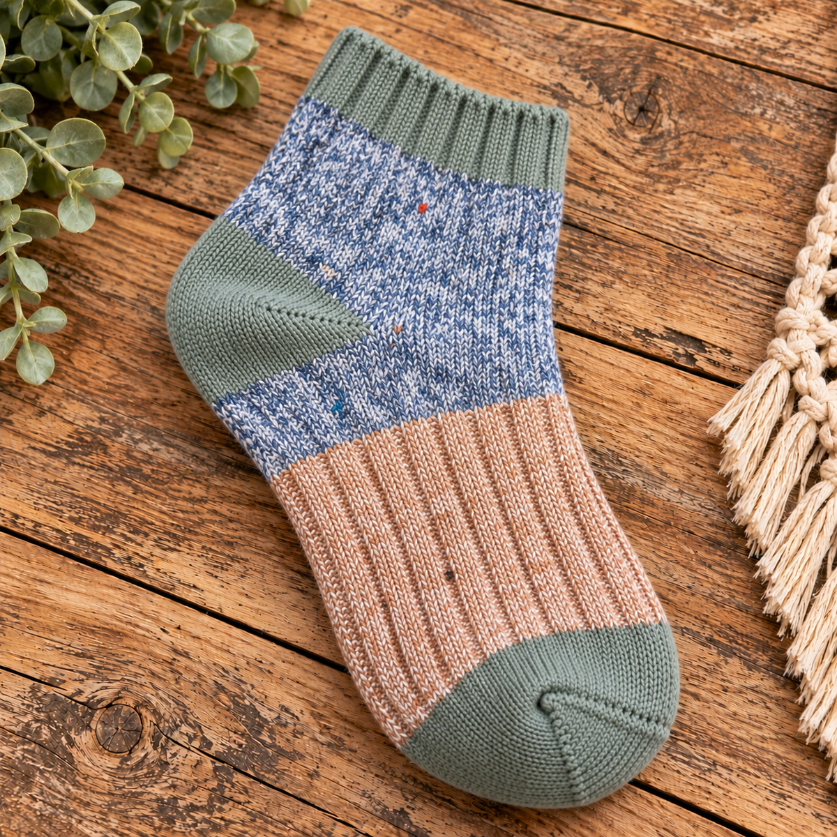 A single hand-knit sock with a color-blocked pattern of sage green, heathered blue, and dusty pink, displayed on a rustic wooden surface with decorative greenery and a macrame fringe.