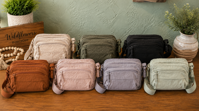 Six linen crossbody bags in different muted colors are lined up on a wooden table against a textured green wall, with decorative plants and a wooden sign in the background.