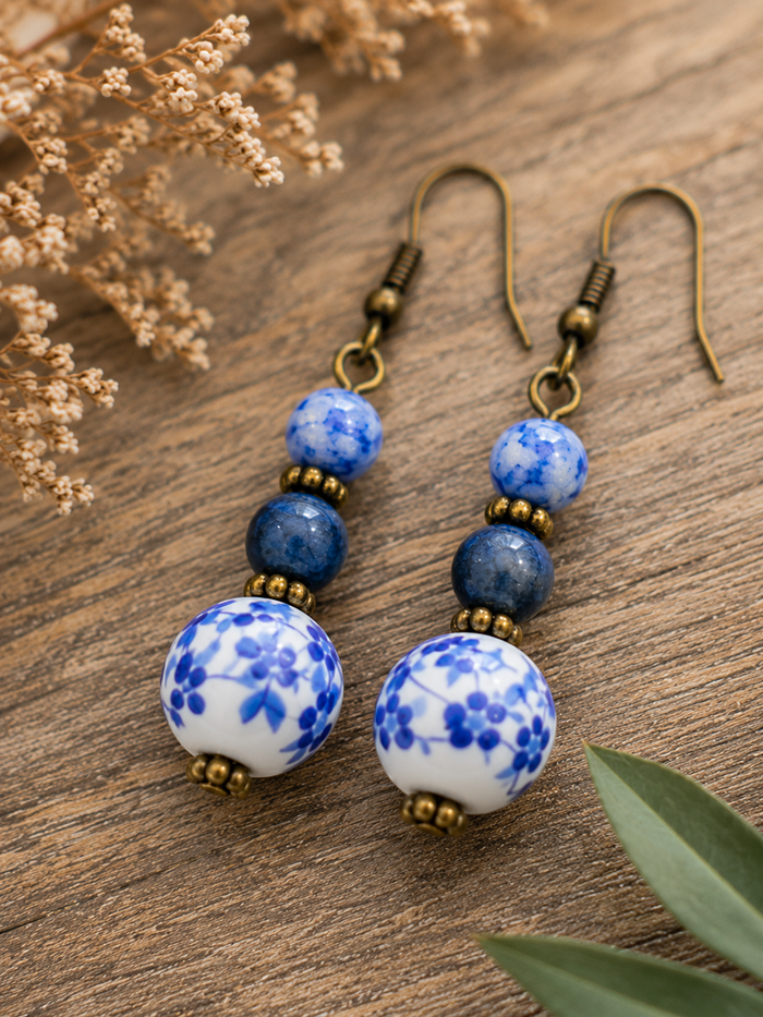 A pair of dangle earrings with blue and white floral ceramic beads, dark blue stone beads, and antique bronze metal hooks, displayed on a rustic wooden surface with dried flowers and green leaves.