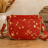 A red shoulder bag with a gold speckled pattern, featuring a gold-trimmed flap and matching patterned strap, displayed on a wooden surface with rustic decorative props in the background.