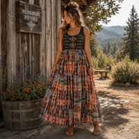 A woman stands outdoors in front of a rustic wooden building, wearing a sleeveless boho maxi dress with a black embroidered bodice and a colorful patchwork skirt in orange, red, blue, and beige tones. A wooden sign on the wall reads 'WILDFLOWERS BOUTIQUE'.