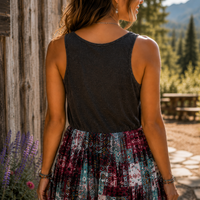 A woman shown from behind wearing a dark charcoal gray sleeveless tank top and a multicolor patchwork print skirt in shades of purple, teal, and white, standing outdoors next to a rustic wooden wall.