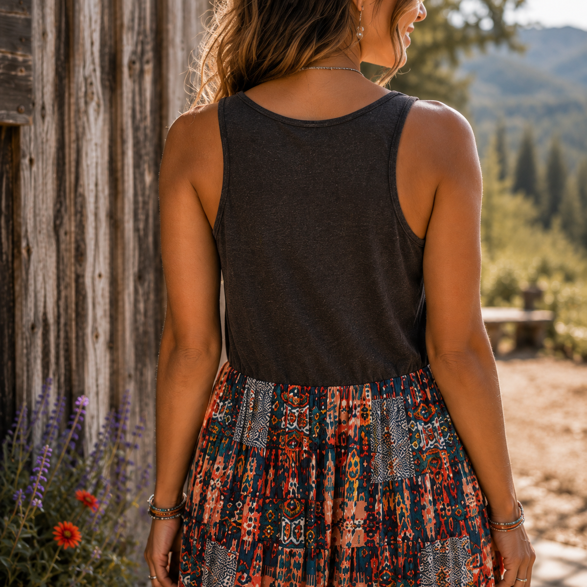 A woman shown from behind wearing a dark charcoal sleeveless tank top and a colorful patterned skirt, standing outdoors next to a rustic wooden wall with a mountain landscape in the background.
