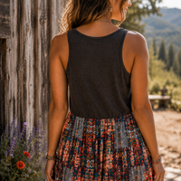 A woman shown from behind wearing a dark charcoal sleeveless tank top and a colorful patterned skirt, standing outdoors next to a rustic wooden wall with a mountain landscape in the background.