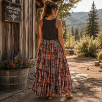 A woman stands outdoors in front of a rustic wooden building, shown from the back. She is wearing a sleeveless black top and a long, pleated maxi skirt with a dense multicolor floral and abstract print in red, orange, blue, and gray tones.
